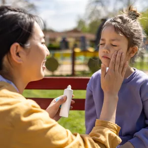 Mom applying sunscreen on child in park.