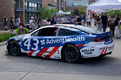 Blue and white race car with AdventHealth branding is displayed at an outdoor community event, with spectators, tents, and storefronts in the background.