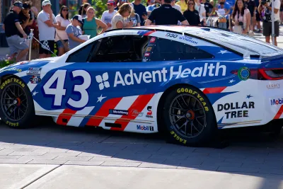 Blue and white race car with AdventHealth branding is displayed at an outdoor community event, with spectators and storefronts in the background.