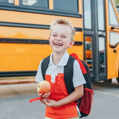A Small Boy Laughs as He Gets Off a School Bus with an Apple in Hand.