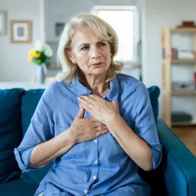 A Woman Sits in Her Living Room With Her Hands on Her Chest, Feeling Her Heartbeat.