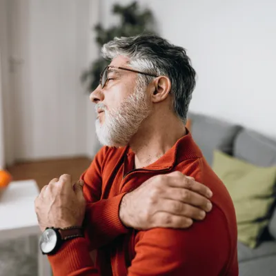 An Older Gentleman Rubs a Sore Shoulder While Sitting on His Sofa at Home