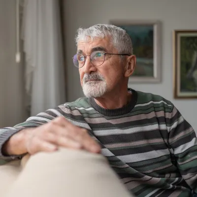 An Senior Man with gray hair and glasses is sitting on a beige couch, wearing a striped sweater.