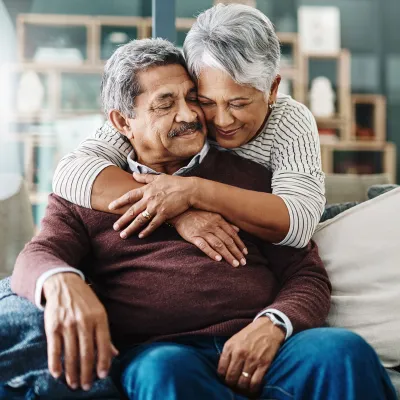 The wife of an old couple hugs her husband on the couch