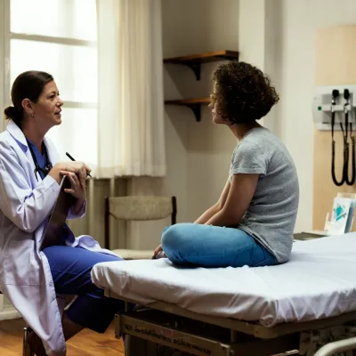 A young woman sits in a hospital room and talks about her health and wellness with her primary care doctor.