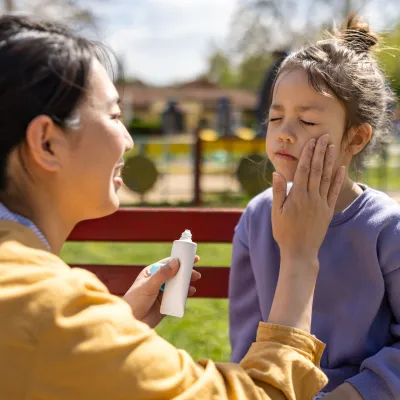 Mom applying sunscreen on child in park.