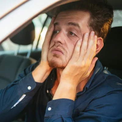 An upset man holds his face with his hands while sitting in a car.