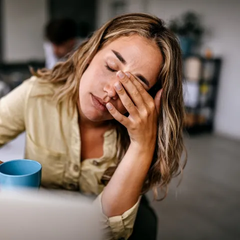 A Woman Presses Her Fingers to the Bridge of Her Nose While Drinking a Cup of Coffee at Home
