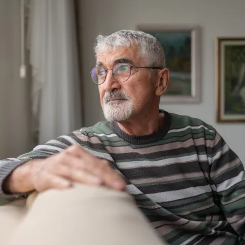 An Senior Man with gray hair and glasses is sitting on a beige couch, wearing a striped sweater.