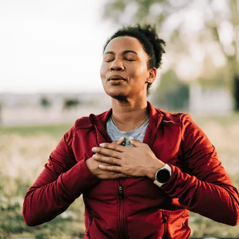 Woman in a red jacket practicing deep breathing outdoors, eyes closed and hands on chest.