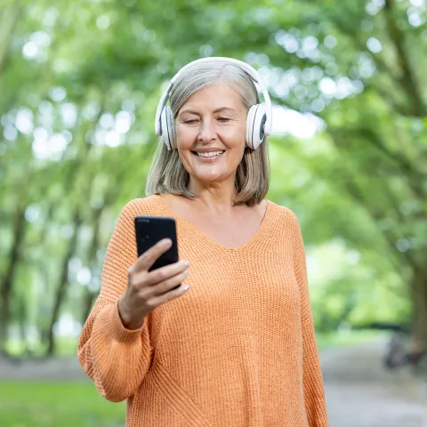 Older woman with headphones on looking at her phone outside.