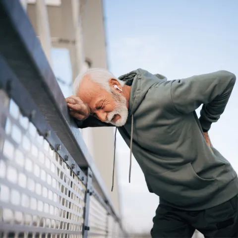 A senior man hunching over against a fence with his hand resting on his back in pain.