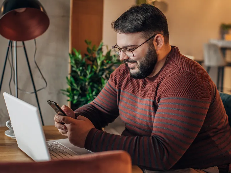 A Man Smiles as He Uses a Cellphone While Sitting at a Desk in front of a Laptop.