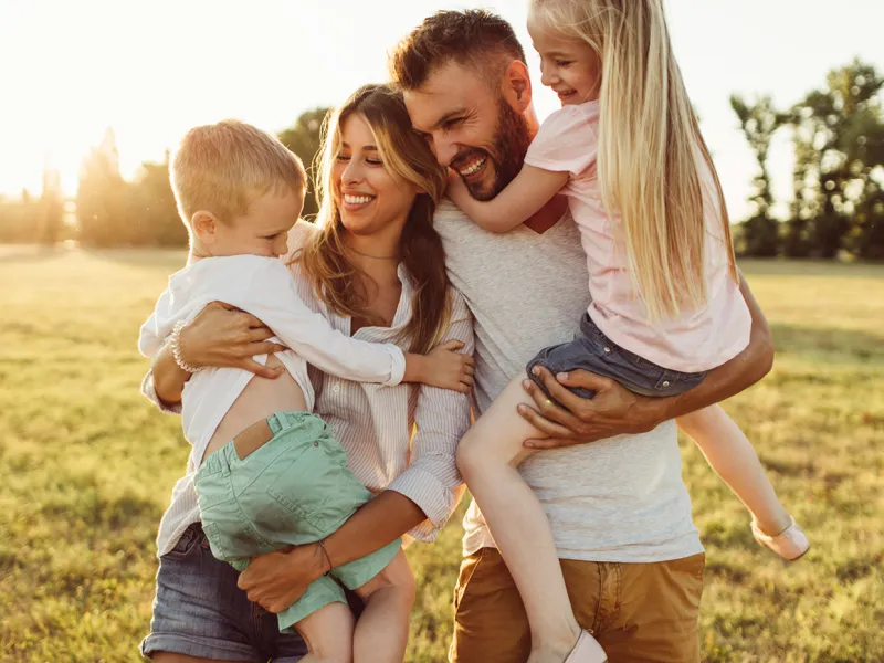 A family of four, the mother holding the son and the father holding the daughter, all laughing and smiling. The sun setting behind them.