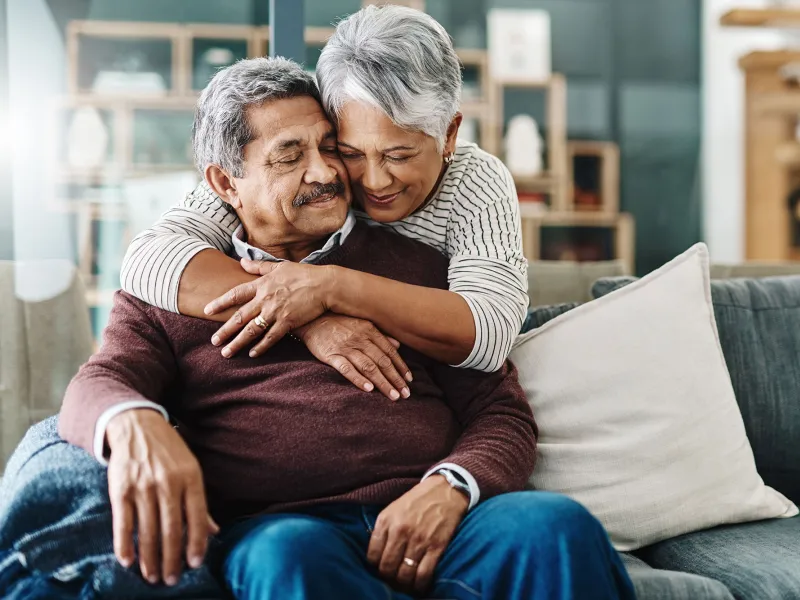 The wife of an old couple hugs her husband on the couch