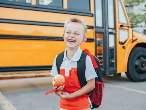 A Small Boy Laughs as He Gets Off a School Bus with an Apple in Hand.