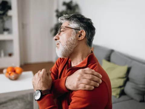 An Older Gentleman Rubs a Sore Shoulder While Sitting on His Sofa at Home