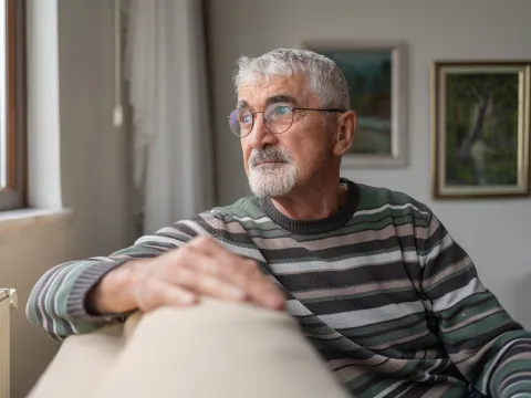 An Senior Man with gray hair and glasses is sitting on a beige couch, wearing a striped sweater.