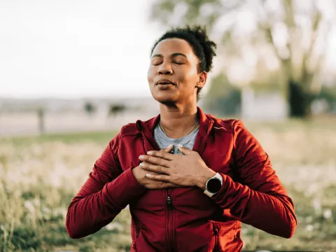 Woman in a red jacket practicing deep breathing outdoors, eyes closed and hands on chest.