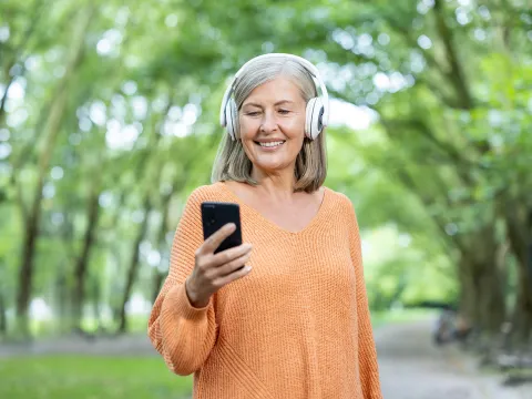 Older woman with headphones on looking at her phone outside.