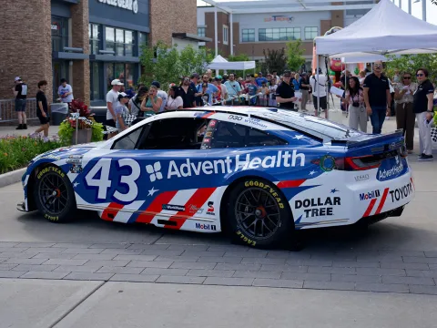 Blue and white race car with AdventHealth branding is displayed at an outdoor community event, with spectators, tents, and storefronts in the background.