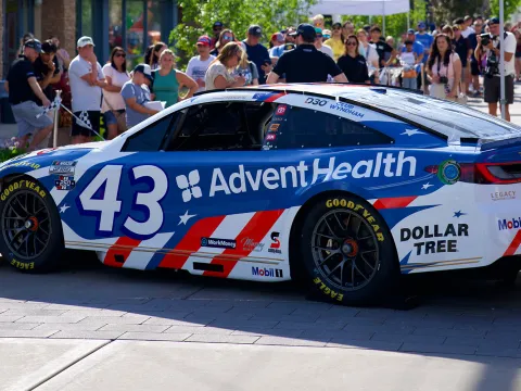 Blue and white race car with AdventHealth branding is displayed at an outdoor community event, with spectators and storefronts in the background.