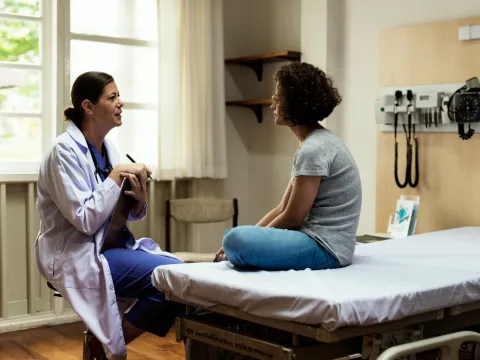 A young woman sits in a hospital room and talks about her health and wellness with her primary care doctor.