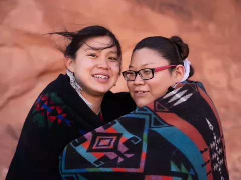 Native American mother and daughter wrapped in a blanket together while outdoors.