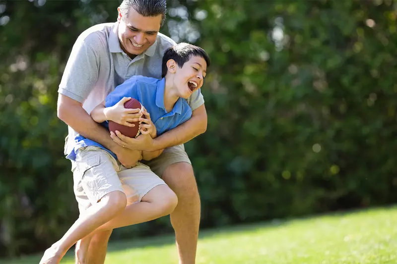 A father playfully tackles his son as they play football outside.
