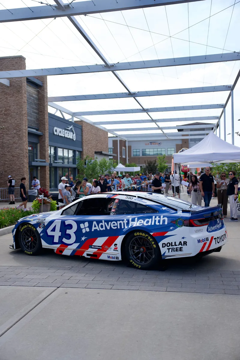 Blue and white race car with AdventHealth branding is displayed at an outdoor community event, with spectators, tents, and storefronts in the background.
