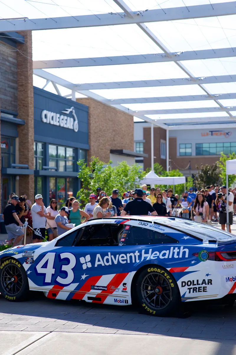Blue and white race car with AdventHealth branding is displayed at an outdoor community event, with spectators and storefronts in the background.